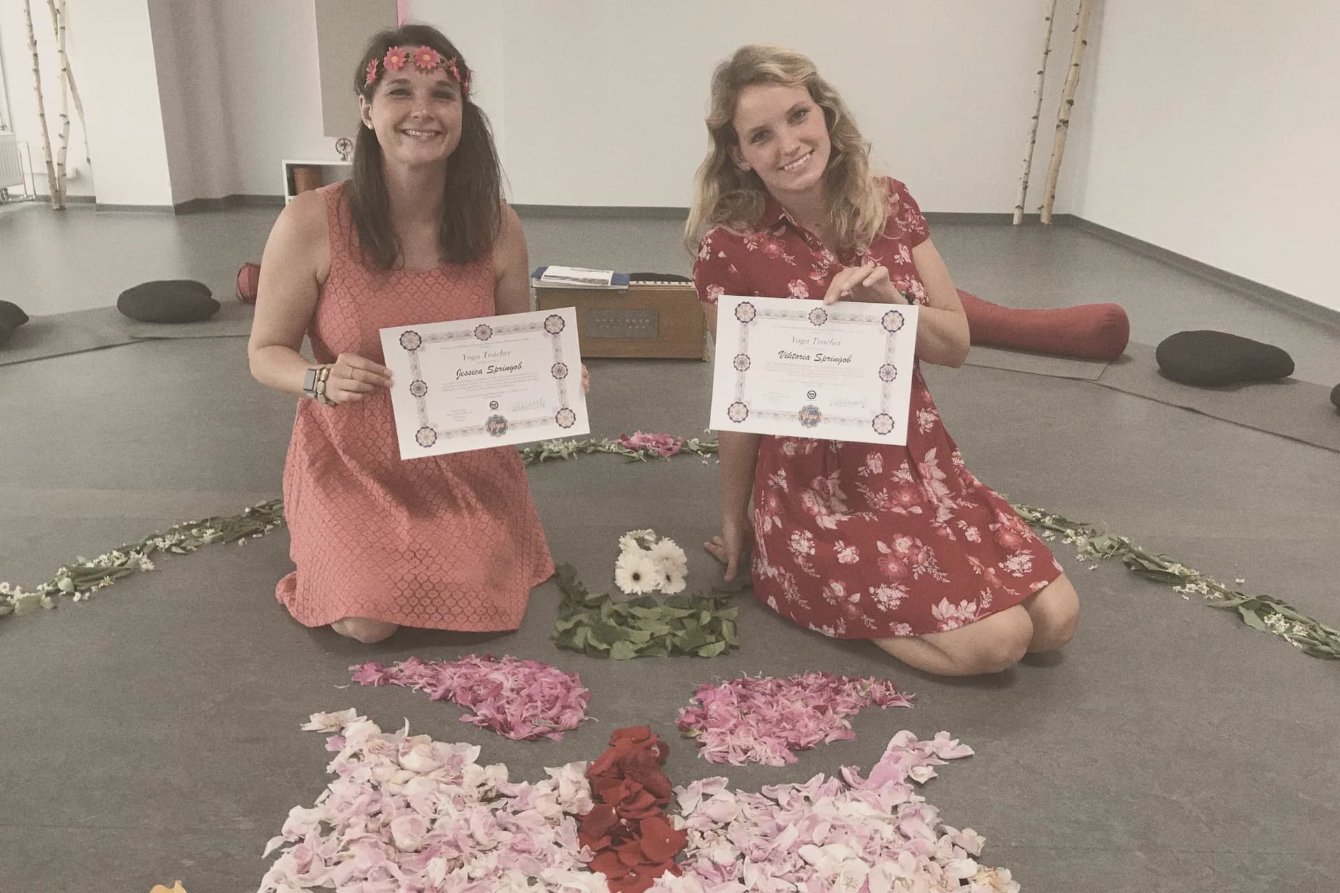 Jessica und Viktoria Springob halten ihre Yoga-Teacher-Zertifikate, vor ihnen ein Mandala aus Blüten